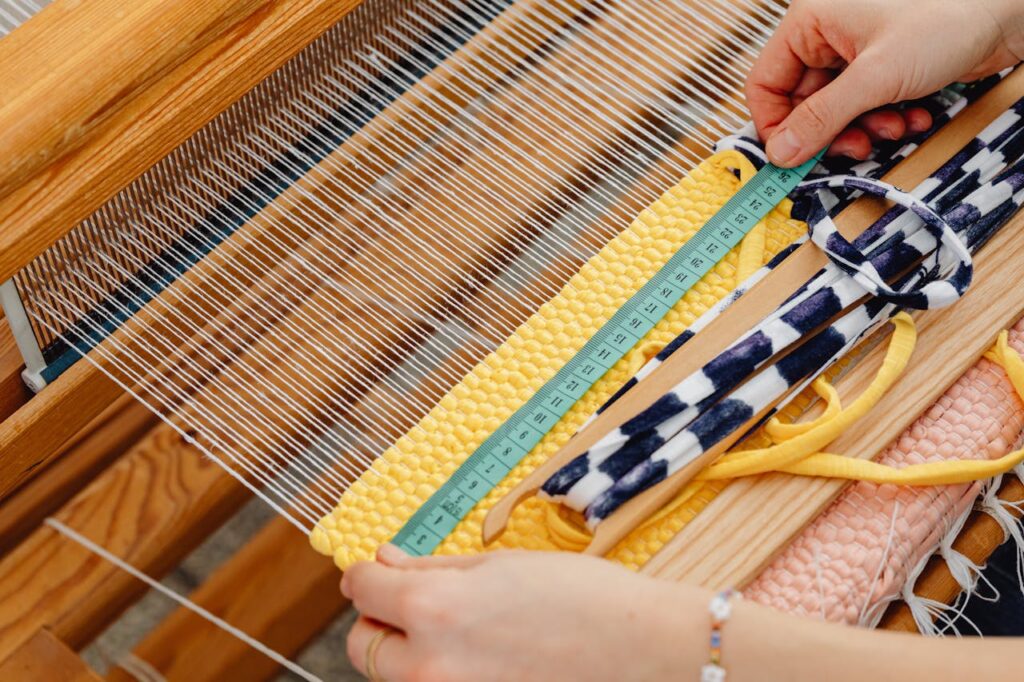 Close-up of hands weaving on a wooden loom with colorful threads and a measuring tape.