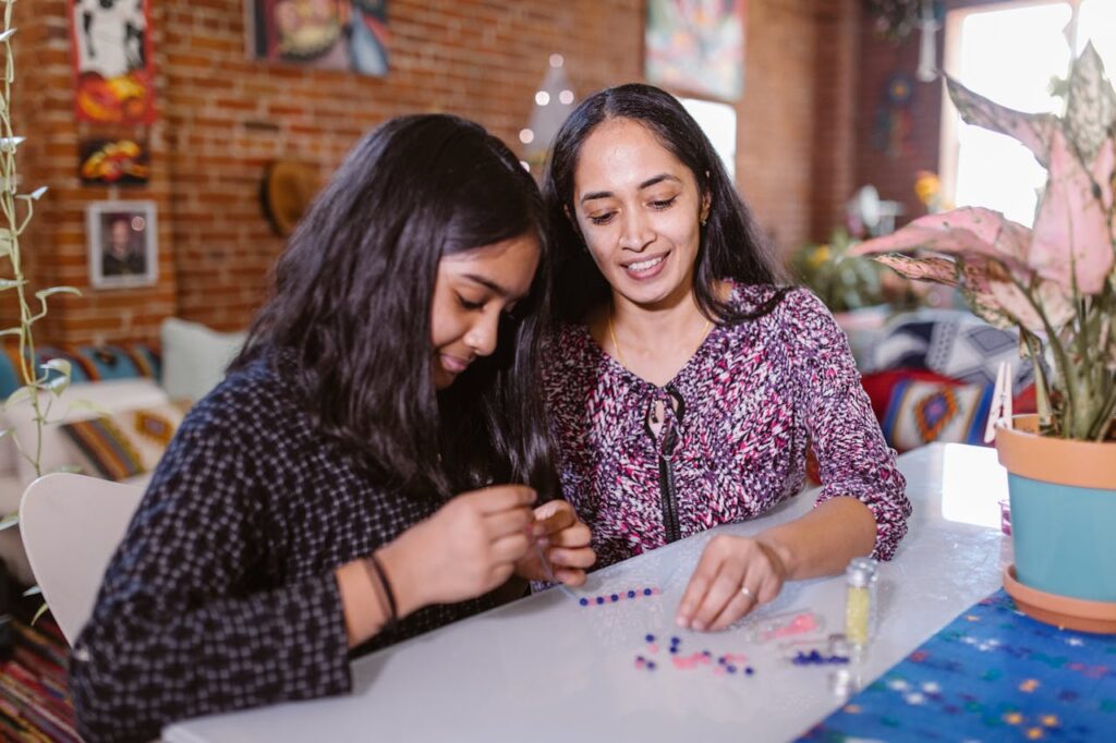 A mother and daughter enjoy crafting beaded jewelry together in a cozy home environment.